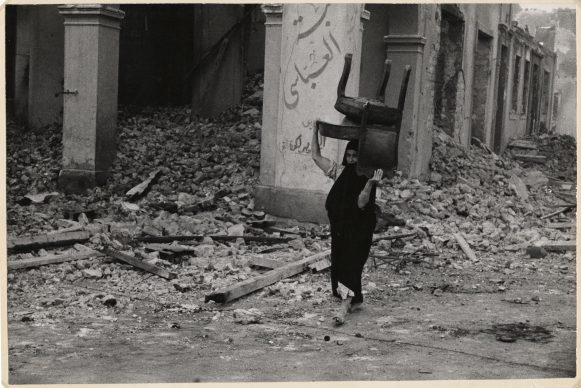 Vrouw loopt door het puin met een stoel op haar hoofd, Port Said, Egypte, november 1956 © Chim (David Seymour),  Magnum  Photos. Courtesy Chim  Estate