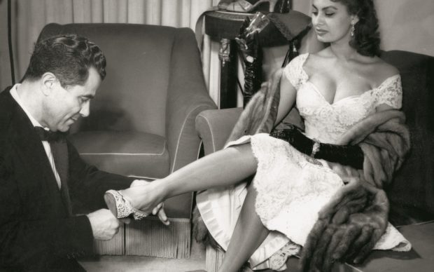 Sophia Loren e Salvatore Ferragamo fotografati insieme durante la serata presso l'Open Gate Club di Roma, organizzata per celebrare il suo nuovo brevetto della pelle di leopardo marino, 28 febbraio 1955. Ferragamo prova una scarpa in merletto all'attrice. © Museo Salvatore Ferragamo